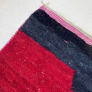 May include: Close-up of a colorful rug with a geometric design. The rug features a vibrant red section, followed by dark blue and black stripes. The edge has a pink and beige striped border.