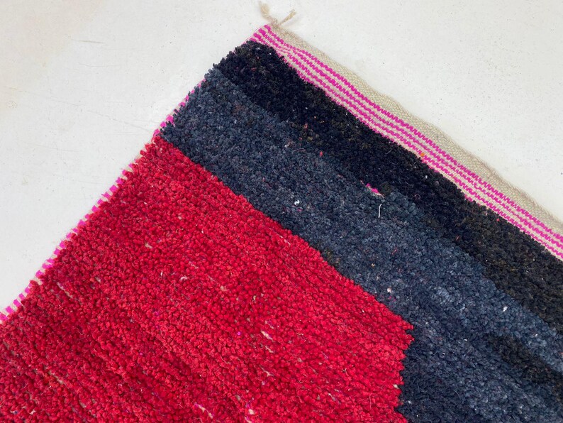 May include: Close-up of a colorful rug with a geometric design. The rug features a vibrant red section, followed by dark blue and black stripes. The edge has a pink and beige striped border.