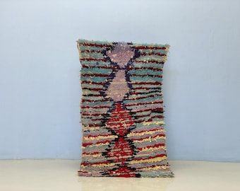 Può includere: Un tappeto rettangolare fatto a mano con un design geometrico. Il tappeto presenta un motivo a diamante centrale in rosso, viola e nero, circondato da strisce orizzontali in vari colori, tra cui blu, giallo e grigio. Il tappeto è lungo circa 61 cm e largo 41 cm.