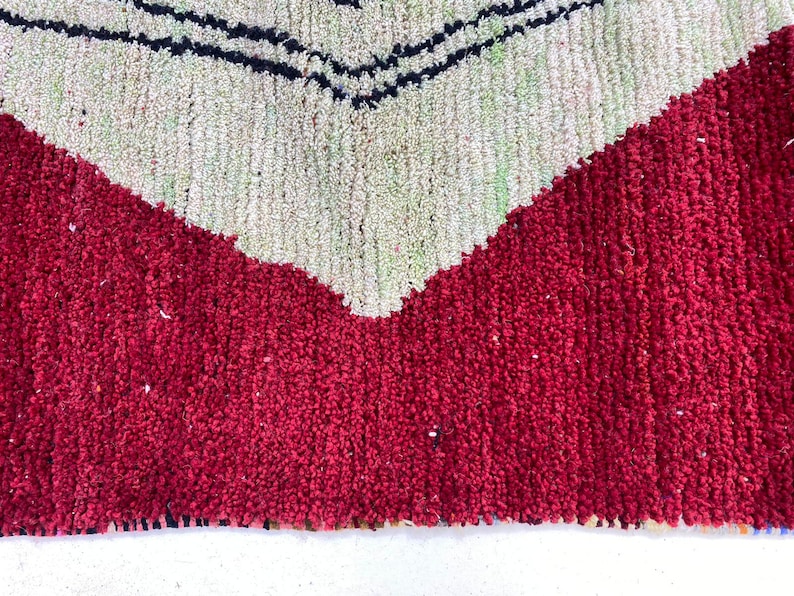 May include: Close-up of a textured rug with a bold geometric design. The rug features a vibrant red base with a large, off-white chevron pattern. Thin black lines accent the chevron, creating a striking contrast. The rug's texture suggests a soft, comfortable feel.