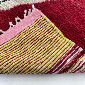 May include: A close-up of a red and yellow striped rug with a pink border. The rug has a textured surface and a folded corner, revealing the underside with yellow and white stripes. White fringe is visible.