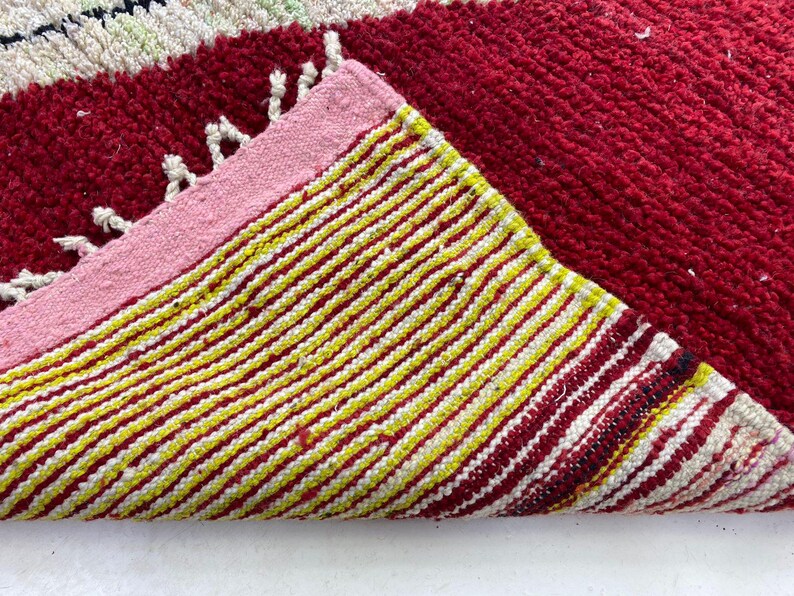 May include: A close-up of a red and yellow striped rug with a pink border. The rug has a textured surface and a folded corner, revealing the underside with yellow and white stripes. White fringe is visible.