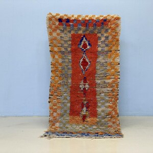 Peut inclure: Tapis rectangulaire tissé avec un panneau central orange orné de motifs en losange rouges, blancs et bleus. Le tapis a une bordure texturée et frangée dans des tons orange, bleu et gris, créant une esthétique rustique.