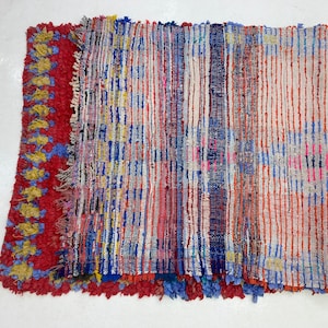 a vibrant, multicolored textile with a striking pattern of vertical stripes in various colors, including red, blue, yellow, and white. The textile appears to be a handmade piece, possibly a rug or a textile art piece, with a textured surface that adds depth and visual interest to the overall composition.