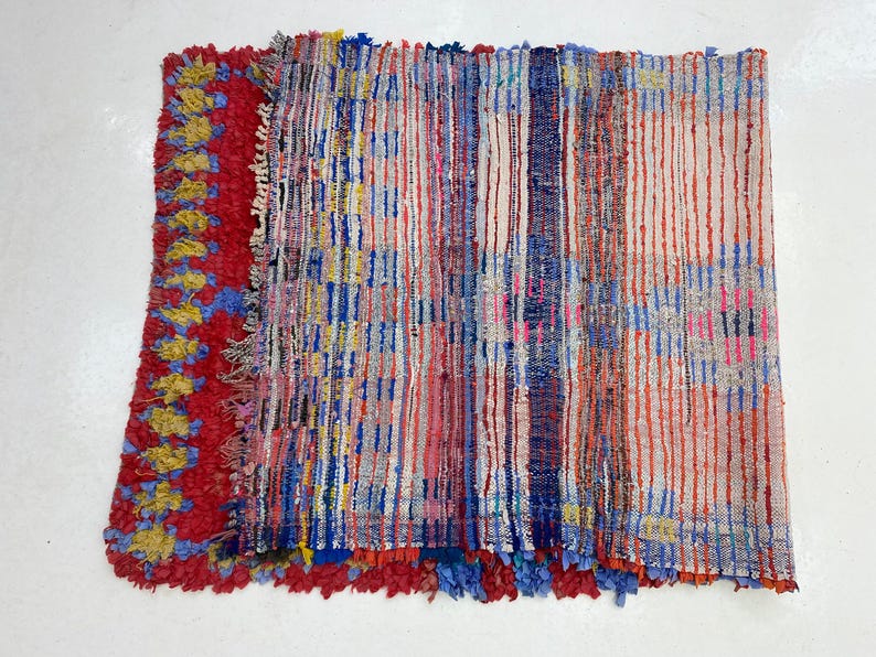 a vibrant, multicolored textile with a striking pattern of vertical stripes in various colors, including red, blue, yellow, and white. The textile appears to be a handmade piece, possibly a rug or a textile art piece, with a textured surface that adds depth and visual interest to the overall composition.
