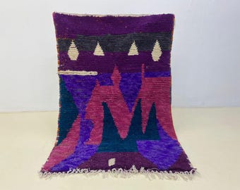 May include: A vibrant, hand-woven rug with a geometric design. The rug features a rich color palette of purple, pink, teal, and cream. The design includes triangles and other shapes. The rug has a fringed edge and is likely made of wool.