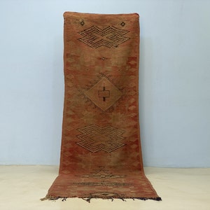 May include: A long, rectangular rug with a reddish-brown and tan color scheme. The rug features geometric patterns in black, including diamond and zigzag shapes. The rug is made of woven material and has a slightly distressed appearance.