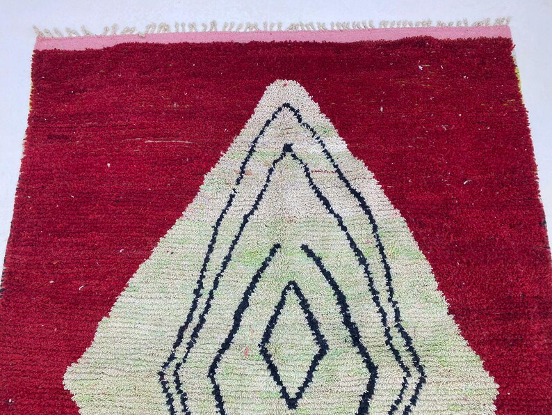 May include: A red and white geometric rug with a diamond pattern. The rug has a red background with a large white diamond shape in the center, outlined in black. The diamond has a green center.