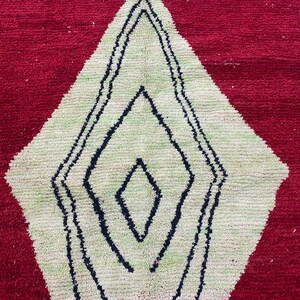 May include: A red rug with a large diamond pattern in the center. The diamond is light green with a black outline and a smaller black diamond in the center. The rug has a textured, woven appearance.
