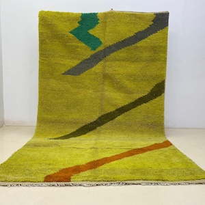 May include: A handmade, rectangular rug with a yellow-green background. The rug features diagonal stripes in gray, dark green, and orange. A zig-zag pattern in green is also present. The rug is made of a thick, textured material.