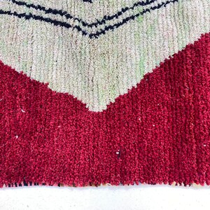 May include: Close-up of a textured rug with a bold geometric design. The rug features a vibrant red base with a large, off-white chevron pattern. Thin black lines accent the chevron, creating a striking contrast. The rug's texture suggests a handmade quality.