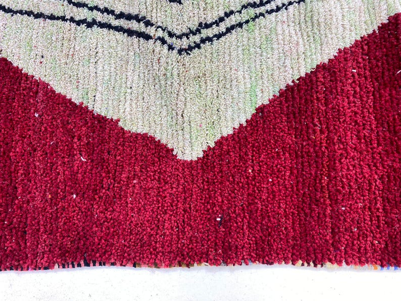 May include: Close-up of a textured rug with a bold geometric design. The rug features a vibrant red base with a large, off-white chevron pattern. Thin black lines accent the chevron, creating a striking contrast. The rug's texture suggests a handmade quality.