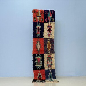 Peut inclure: Un tapis rectangulaire à motifs géométriques répétitifs. Le tapis présente un motif patchwork avec des carrés rouges, bleu marine, crème et violets. Chaque carré contient un motif abstrait différent de différentes couleurs.