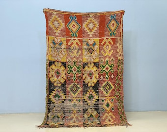 Puede incluir: Una alfombra rectangular tejida a mano con un patrón geométrico. La alfombra presenta una paleta de colores rojo, amarillo, verde y negro. El diseño incluye motivos de diamantes y florales. La alfombra tiene un borde con flecos y se exhibe contra una pared azul claro.