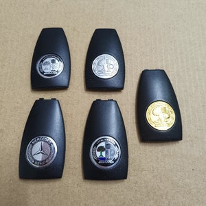 Peut inclure: Cinq porte-clés noirs avec différents logos argentés et dorés. Les logos comprennent Mercedes-Benz, Affalterbach et AMG.