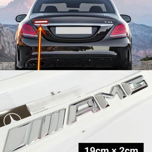 Puede incluir: Emblema cromado "AMG", 19 cm x 2 cm, para coche. El emblema se muestra sobre una superficie blanca con un coche negro al fondo. El coche es negro con luces traseras rojas. También se muestra el modelo del coche, C43.