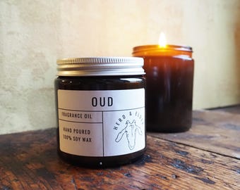 May include: A brown glass candle jar with a silver lid. The label reads "OUD FRAGRANCE OIL HAND POURED 100% SOY WAX" and features a black and white illustration of a horse's head. The candle is sitting on a wooden surface.