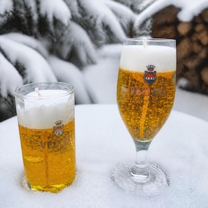 Könnte beinhalten: Zwei bierförmige Kerzen in klaren Glasbehältern. Links ein zylindrisches Glas, rechts ein Stielglas. Beide haben eine goldgelbe Flüssigkeitsbasis, eine weiße Schaumkrone, einen zentralen Docht und ein Wappenlogo mit dem Schriftzug VELTINS.