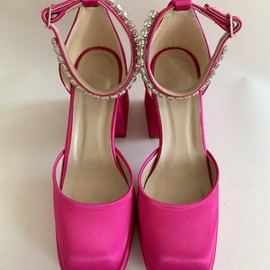 May include: A pair of bright pink satin heeled shoes with a square toe and ankle strap adorned with rhinestones.