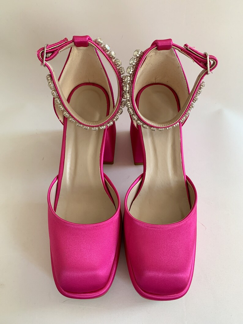 May include: A pair of bright pink satin heeled shoes with a square toe and ankle strap adorned with rhinestones.