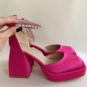 May include: Pink satin platform heels with a rhinestone ankle strap. The heels have a chunky square shape.