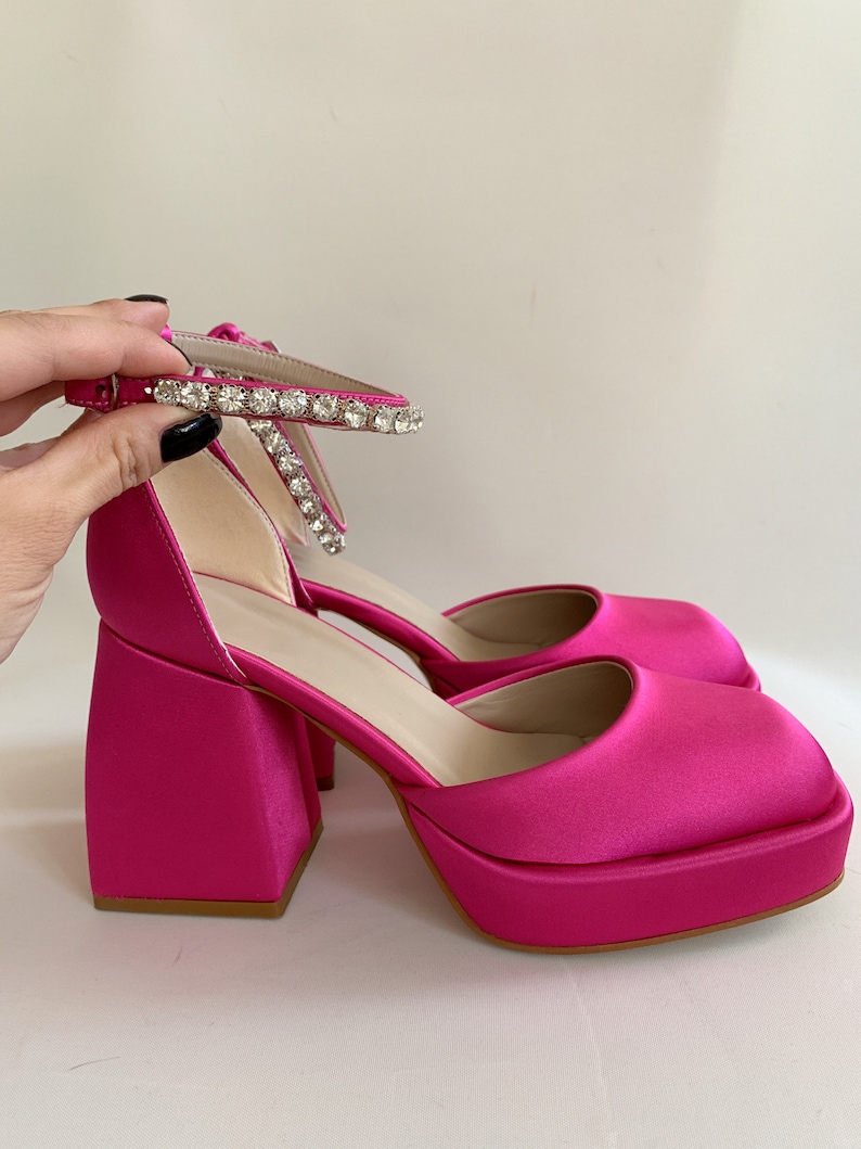May include: Pink satin platform heels with a rhinestone ankle strap. The heels have a chunky square shape.