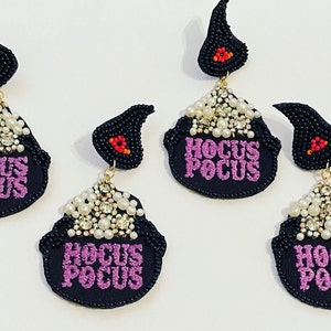 May include: A pair of black beaded earrings with a cauldron charm. The cauldron is filled with white pearls and rhinestones and has the words "Hocus Pocus" embroidered in purple.
