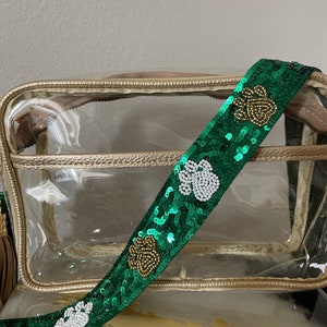 May include: Clear plastic tote bag with a gold trim and a green and white sequined shoulder strap with shamrock designs. "MADE IN" is visible on the bag.