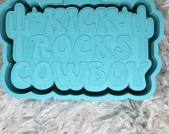 May include: A light blue silicone mold for making resin crafts. The mold features the text "Kick Rocks Cowboy" with a cactus on each side.
