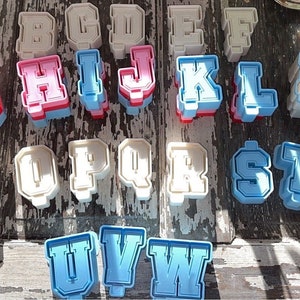 May include: A set of 26 alphabet cookie cutters in white, pink, and blue. The cutters are shaped like block letters with a white outline and a contrasting color inside.