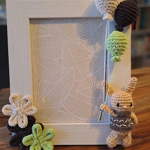 Può includere: Una cornice in legno bianco decorata con elementi all'uncinetto. La cornice è adornata da tre palloncini all'uncinetto beige, marroni e verdi, tenuti da un coniglietto all'uncinetto. Fiori all'uncinetto beige, marroni e verdi sono posizionati in basso.