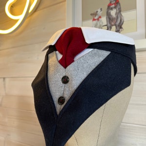 May include: A navy blue, gray, and red dog bandana with a tuxedo design. The bandana has two black buttons and a white collar.