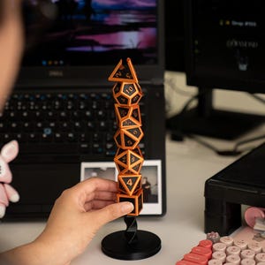 Könnte beinhalten: Ein Turm aus orangefarbenen und schwarzen polyedrischen Würfeln auf einem schwarzen Ständer. Die Würfel sind gestapelt und zeigen ihre Zahlen. Die Würfel befinden sich vor einem Laptop und einem Computermonitor.