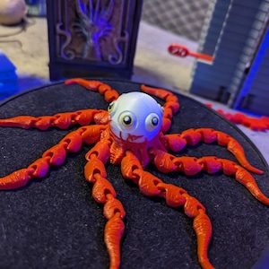 Peut inclure: Figurine d'un poulpe orange imprimé en 3D, avec une tête blanche et des yeux de dessin animé. Le poulpe a des tentacules articulés et est posé sur une surface sombre. La figurine a un design fantaisiste et ludique.
