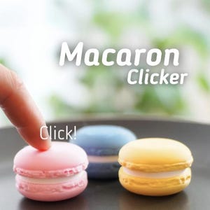 May include: Three colorful macarons, pink, blue, and yellow, are displayed on a dark plate. The word "Macaron Clicker" is written above the macarons, with the word "Click!" next to the pink macaron.