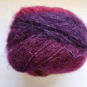 Pu&ograve; includere: Un gomitolo di filato nei toni del viola e magenta, con fili bianchi. Il filato ha una consistenza morbida e soffice, e i colori si fondono. Adatto per progetti di maglia o uncinetto.