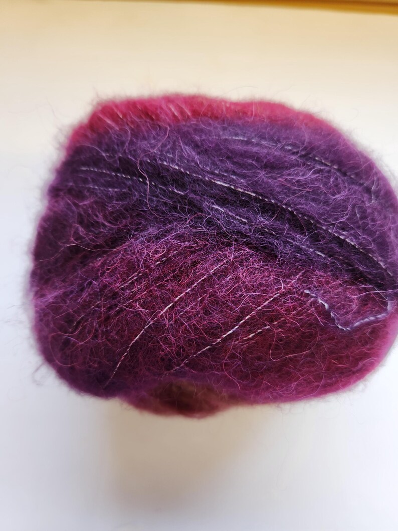 Pu&ograve; includere: Un gomitolo di filato nei toni del viola e magenta, con fili bianchi. Il filato ha una consistenza morbida e soffice, e i colori si fondono. Adatto per progetti di maglia o uncinetto.