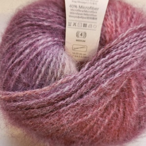 May include: A ball of pink and purple yarn with a fluffy texture. The label shows the yarn composition: 60% Kid Mohair and 40% Microfibre. The label also indicates a medium weight. The yarn is suitable for knitting or crochet projects.