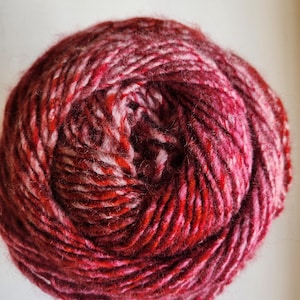 Könnte beinhalten: Ein Knäuel Garn in Rot-, Rosa- und Weißtönen. Das Garn scheint eine dicke, strukturierte Wolle zu sein, perfekt für Strick- oder Häkelprojekte. Die Farben verschmelzen und erzeugen ein optisch ansprechendes und lebendiges Aussehen.