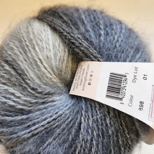 Puede incluir: Una madeja de hilo gris y blanco con una etiqueta adjunta. La etiqueta dice "MADE IN ITALY FOR KNITTING FEVER" e incluye un código de barras y el número de color 898. El hilo parece suave y esponjoso, ideal para proyectos de tejido o ganchillo.