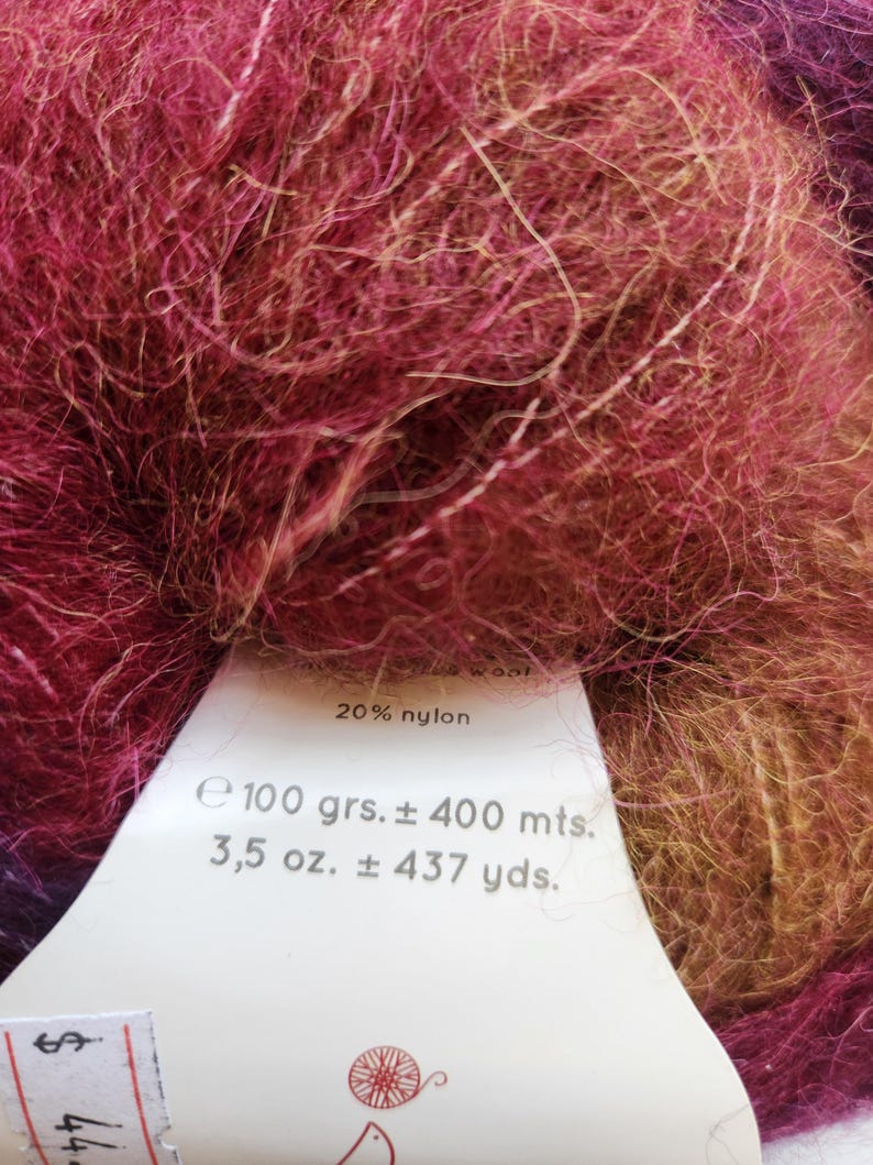 Pu&ograve; includere: Primo piano di un gomitolo di filato nei toni del bordeaux, rosa e oro. Il filato ha un aspetto soffice e strutturato. Un'etichetta indica che il filato &egrave; 20% nylon, 100 grammi e circa 400 metri. Ideale per progetti di maglia e uncinetto.