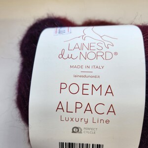 Pu&ograve; includere: Primo piano di un gomitolo di filato bordeaux scuro con un'etichetta. L'etichetta recita "LAINES du NORD" e "POEMA ALPACA Luxury Line". Il filato &egrave; prodotto in Italia e ha un codice a barre.