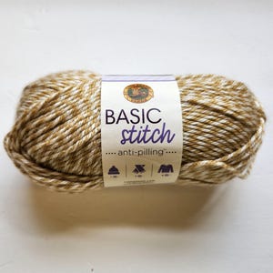 May include: A skein of Basic Stitch anti-pilling yarn in a blend of tan and white. The label features the brand logo and the words "BASIC stitch" in purple. Icons for a hat, scarf, and sweater are also visible.