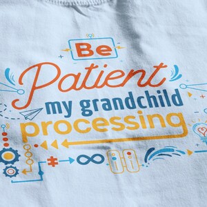May include: Three folded t-shirts with a light blue, white, and beige color scheme. The shirts feature a graphic design with the text "Be Patient my grandchild processing" in orange and blue.