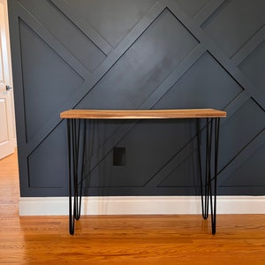 May include: A wooden console table with a natural wood finish and black hairpin legs. The table is in front of a dark grey wall with a geometric pattern.