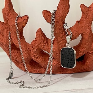 Peut inclure: Un collier argenté avec un pendentif rectangulaire noir. Le pendentif a une surface texturée et est serti dans un cadre argenté. La chaîne du collier est également argentée.