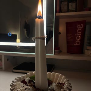 May include: A white taper candle with a lit flame, tied with a thin string, standing upright in a white, scalloped-edge ceramic bowl filled with decorative stones, dried petals, and small white fabric pieces, creating a centerpiece arrangement.