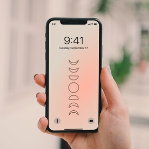 May include: A black smartphone with a white screen displaying the time 9:41 and the date Tuesday, September 17. The background of the screen is a gradient of pink and orange. A stylized image of the phases of the moon is displayed on the screen.