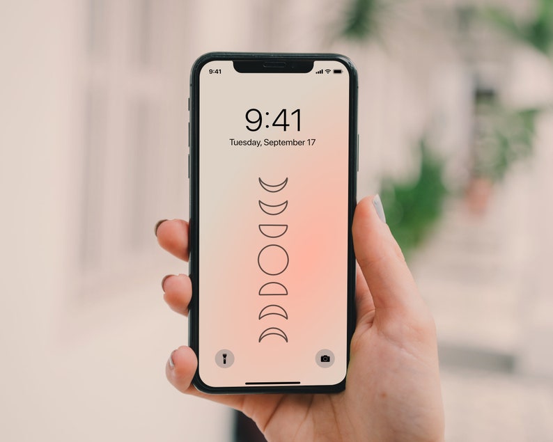 May include: A black smartphone with a white screen displaying the time 9:41 and the date Tuesday, September 17. The background of the screen is a gradient of pink and orange. A stylized image of the phases of the moon is displayed on the screen.
