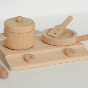May include: A wooden toy kitchen set with a pot, pan, spatula, and spoon. The set is made of natural wood and has a light brown color.
